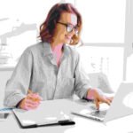 Happy brunette businesswoman in glasses using laptop computer while working in light apartment